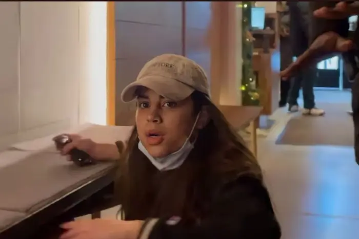 A screenshot of the woman wanted by NYPD for scratching and attacking a Black man and his teenage son at the Arlo Hotel. She is wearing a tan baseball cap and a mask pulled down.